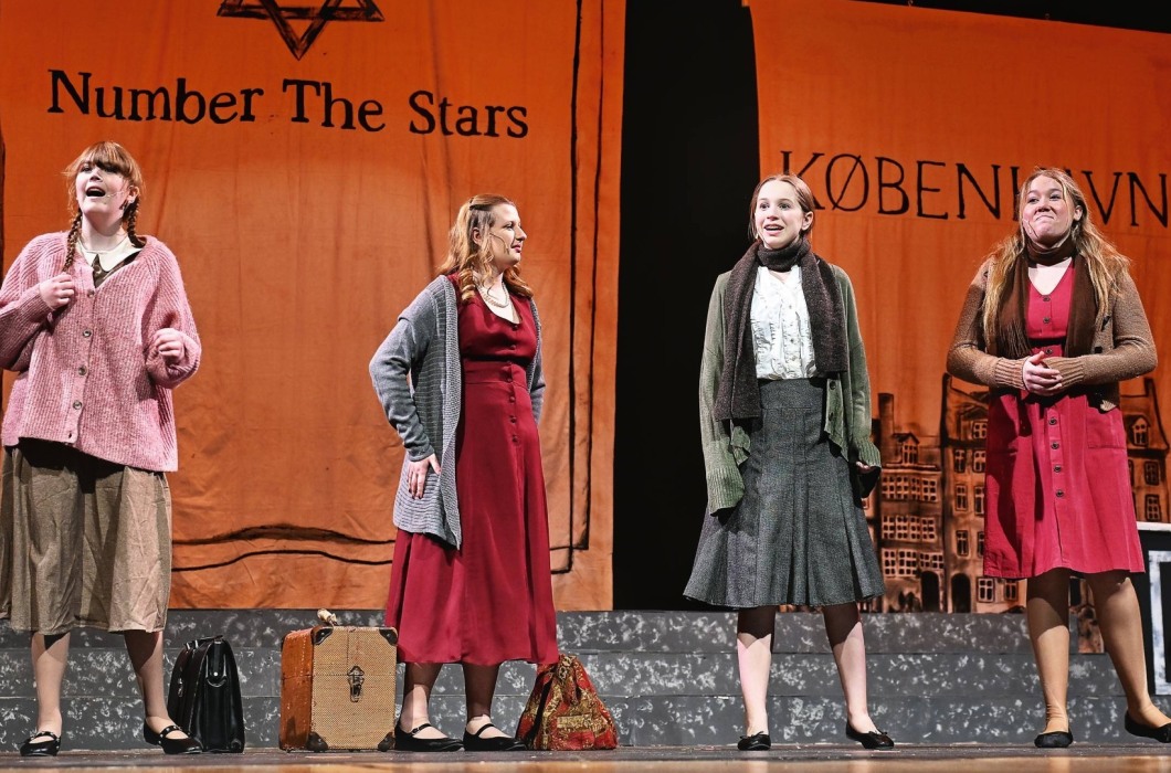 Four actors stand on a stage with backdrops for the show Number the Stars behind them. 